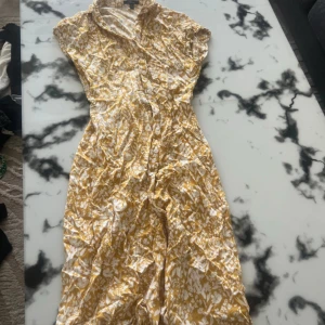 Gulvit blommig kortklänning - Säljer en söt gulvit kortklänning med blommigt mönster och knappar framtill. Klänningen har krage och korta ärmar, perfekt för varma dagar. Materialet känns lätt och luftigt, vilket gör den superbekväm att bära.