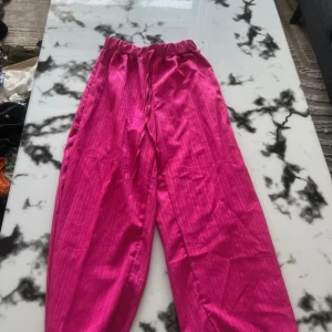 Rosa vida byxor med snörning - Snygga vida byxor i starkt rosa färg med resår och snörning i midjan. Lätta och luftiga, perfekta för varmare dagar. Materialet har en lätt glans och känns mjukt mot huden. Passar dig som vill sticka ut med färg och stil. LINNEBYXOR