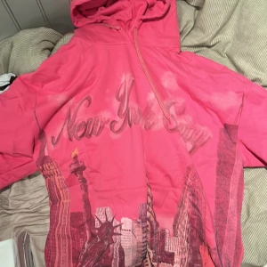 Rosa hoodie med New York City-tryck - Cool rosa hoodie med tryck av New York City och stadssilhuett framtill. Tröjan har huva, dragkedja och långa ärmar. Perfekt för dig som gillar streetstyle och vill sticka ut med färg och motiv.