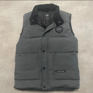  Canada Goose vest  - Snygg grå dunväst från Canada Goose med hög krage, två stora fickor framtill och klassisk logotyp på bröstet. Västen har knappar och dragkedja framtill samt diskret Canada Goose-märke på ena fickan. Perfekt för lager-på-lager-stil.