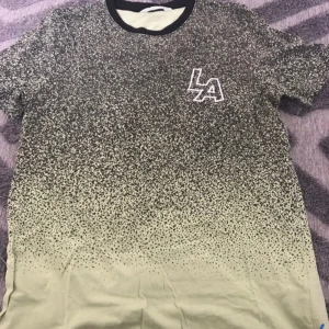 Svart och grön LA t-shirt med mönster - Cool t-shirt med svart och ljusgrön färgton och ett prickigt mönster som tonar ut mot nederkanten. Svart ribbad krage och LA-tryck på bröstet. Perfekt för dig som gillar streetstyle och vill sticka ut lite extra.