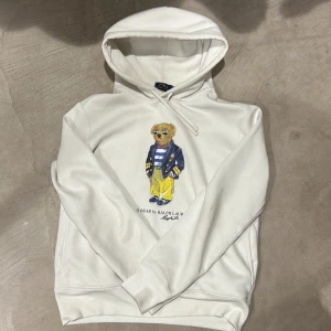 Vit hoodie Polo Bear Ralph Lauren - Vit hoodie från Polo Ralph Lauren med ikoniska Polo Bear-trycket framtill. Tröjan har huva med snörning och är tillverkad i mjuk bomull. Perfekt för dig som gillar streetstyle och klassiska märken.