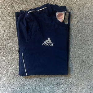 Mörkblå Adidas t-shirt med vita detaljer - Snygg mörkblå t-shirt från Adidas med klassisk logga på bröstet och vita linjer längs axlar och sidor. Perfekt för träning eller chill. Tillverkad i mjuk bomull för skön känsla hela dagen.