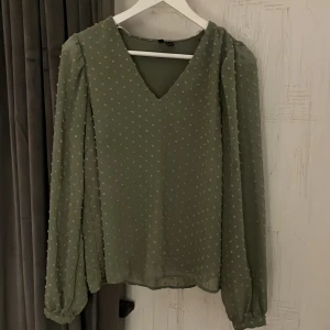 Grön blus med prickar från Vero Moda - Snygg grön blus från Vero Moda med diskreta prickar och v-ringning. Blusen har långa ärmar med lätt puff och är tillverkad i ett tunt, flowy material som ger en luftig känsla. Perfekt att styla med jeans eller kjol för en trendig look. Endast använd 1 gång