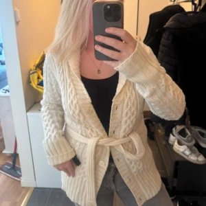 Beige stickad kofta med bälte - Mysig beige stickad kofta med kabelstickat mönster, bred krage och knytskärp i midjan. Långärmad modell med ribbade muddar och knappar framtill. Perfekt för lager-på-lager och chill dagar. Något nopprig och nån tråd som hänger