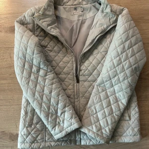Ljusgrå quiltad jacka från greenpoint - Quiltad jacka i ljusgrått från greenpoint med dragkedja framtill och ståkrage. Jackan har ett rutigt mönster och är tillverkad i syntetmaterial. Perfekt för dig som gillar stilren och enkel design med lite edge.