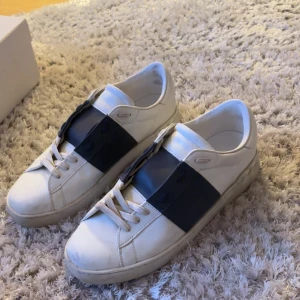 Valentino Garavani Open sneakers vit/blå - Säljer ett par snygga Valentino Garavani Open sneakers i vitt skinn med bred marinblå rem över mitten. Klassisk rund tå, platt sula och vita snören. Kommer med originalkartong och taggar. Perfekt för dig som gillar exklusiva märken och stilren design.          Använda cirka 10 gånger 9/10 skick.      Storlek 45 passar cirka 43/44