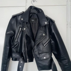 Svart skinnjacka från Bik Bok XS - Snygg svart skinnjacka från Bik Bok i klassisk bikerstil med silvriga dragkedjor, knappar och bälte i midjan. Jackan har flera fickor och coola detaljer som axelklaffar och croppad passform. Perfekt för att lyfta vilken outfit som helst.