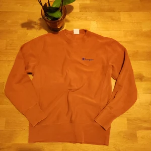 Orange sweatshirt från Champion - Säljer en orange sweatshirt från Champion med broderad logga på bröstet och ärmen. Tröjan har rund halsringning och långa ärmar. Perfekt för dig som gillar streetwear och vill ha en snygg basic i garderoben.