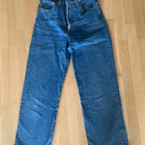 Levi's Ribcage Straight blå jeans - Säljer ett par Levi's Ribcage Straight jeans i klassisk blå denim. Hög midja och raka ben ger en snygg vintagekänsla. Jeansen har fem fickor, knappgylf och tydliga Levi's-detaljer som röd etikett och logoknapp. Perfekta för en avslappnad och trendig look.