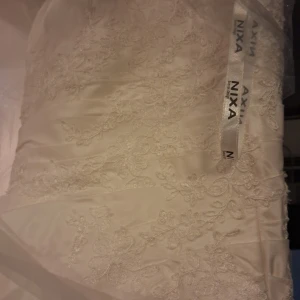 Vit långklänning med spets från Nixa - Elegant vit långklänning från Nixa med vackra broderade spetsdetaljer och blommigt mönster. Klänningen har en klassisk, figurnära passform och romantiska detaljer som ger en lyxig känsla. Perfekt för dig som vill ha något extra speciellt.