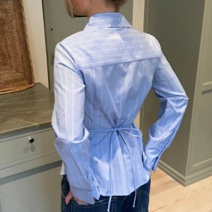 Blå randig skjorta med snörning  - Snygg blå skjorta med vita ränder och figurnära passform från design by Si. Skjortan har långa ärmar, klassisk krage, knappar framtill och snörning i ryggen som ger en cool siluett. Materialet är 53% bomull och 45% polyester, vilket gör den både mjuk och lätt.