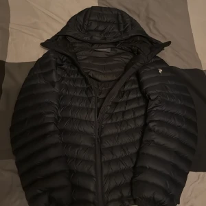 mörkblå dunjacka Peak Performance M - mörkblå dunjacka från Peak Performance i storlek M. Jackan har huva, quiltad design och dragkedja framtill. Perfekt för kyliga dagar och har en clean, sportig look. Passar dig som gillar stilrena och funktionella plagg.