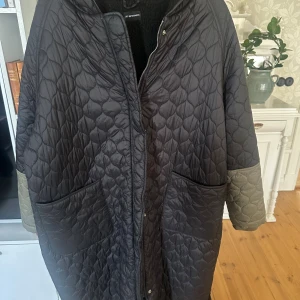 Quiltad svart jacka från M. Wiesneck - Quiltad svart kappa från M. Wiesneck med kontrasterande gröna ärmslut, stora fickor och huva. Jackan har dragkedja och tryckknappar framtill samt en oversized passform. Går ungefär till mitten av vaden/ner till foten. Har fleecefodring inuti.