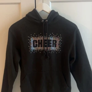 Svart hoodie med glittrig CHEER-text - Svart hoodie med huva och snörning, dekorerad med glittriga paljetter som bildar ordet CHEER på bröstet. Tröjan har en klassisk känguruficka framtill och långa ärmar. Perfekt för dig som håller på med cheerleading. Ordinarie pris 420kr