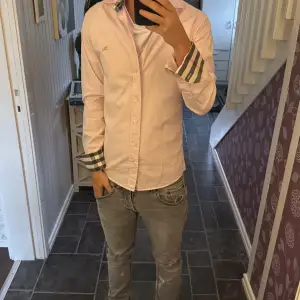 Säljer en ljusrosa skjorta från Burberry med klassiska rutiga detaljer på krage och manschetter. Skjortan har normal passform, långa ärmar och knäppning framtill. Broderad Burberry-logga på bröstet. Perfekt för dig som gillar stilrena och exklusiva plagg. Sitter bra på mig som är 170