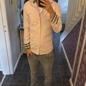 Ljusrosa Burberry skjorta med rutiga detaljer - Säljer en ljusrosa skjorta från Burberry med klassiska rutiga detaljer på krage och manschetter. Skjortan har normal passform, långa ärmar och knäppning framtill. Broderad Burberry-logga på bröstet. Perfekt för dig som gillar stilrena och exklusiva plagg. Sitter bra på mig som är 170