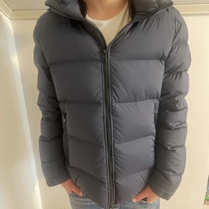 J.Lindeberg jacka  - Snygg mörkgrå dunjacka från J.Lindeberg i storlek L. Jackan har en puffad design, huva och dragkedja framtill. Perfekt för kalla dagar och har en clean look med diskret logga på ärmen. Passformen är normal och jackan är långärmad. Modellen är 181 och väger 64 och den är något stor som man kan se. 