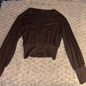 Brun figursydd kofta från Gina Tricot - Snygg brun kofta från Gina Tricot i storlek S. Modellen är croppad med v-ringning, långa ärmar och knappar framtill. Aldrig använt tidigare♥️