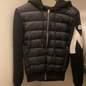Svart Moncler pufferjacka med huva - Säljer en svart pufferjacka från Moncler med huva och dragkedja framtill. Jackan har stickade ärmar, quiltad front och en stor vit detalj på ena ärmen. Snygg och sportig look med snörning i huvan och två fickor framtill.