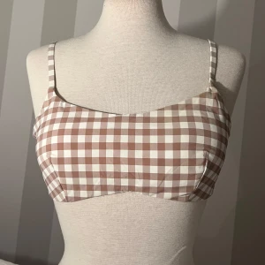 Brun och vit rutig bikini - Söt bikini med brunt och vitt rutigt mönster. Toppen har smala axelband och rak skärning framtill, med spänne i ryggen. Bikinitrosorna har klassisk passform och samma rutiga design.