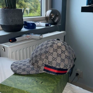 Gucci keps navy - Säljer en snygg Gucci keps i beige och marinblått med klassiskt GG-monogram över hela kepsen. Kepsen har en röd och marinblå rand på sidan och justerbar rem baktill. Materialet är bomullscanvas och insidan är fodrad i svart tyg. Perfekt för dig som vill sticka ut.