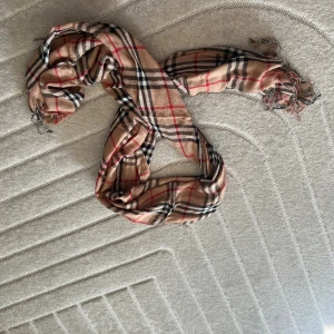 Beige rutig halsduk med fransar - Snygg beige halsduk med klassiskt rutigt mönster i svart, vitt och rött. Lätt och tunn i materialet, perfekt att slänga över axlarna. Fransar längs kanterna ger en chill vibe. Passar till de flesta outfits och är superenkel att styla.