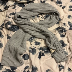 Ljusgrå ribbstickad halsduk East West - Snygg och mjuk ljusgrå halsduk från East West med ribbstickad struktur. Perfekt att vira flera varv runt halsen för en stilren look. Klassisk design som passar till de flesta jackor och outfits.