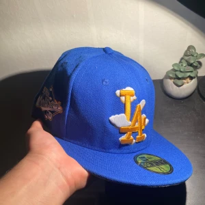 La Dodgers keps New Era - Säljer en blå LA Dodgers keps från New Era 59FIFTY med broderad LA-logga i gult och vitt med molndetaljer. På sidan finns ett broderat 1988 World Series-märke. Kepsen har platt skärm i grått och klassisk passform. Perfekt för dig som gillar sportig streetstyle.
