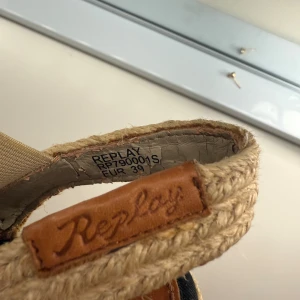 Replay sandaler med flätad detalj - Snygga sandaler från Replay med flätad beige hälrem och svarta korslagda band framtill. Sulan har en naturfärgad, rep-liknande yta och insidan är märkt med Replay-loggan i brunt skinn. Perfekt för dig som gillar chill och stilren sommarlook.