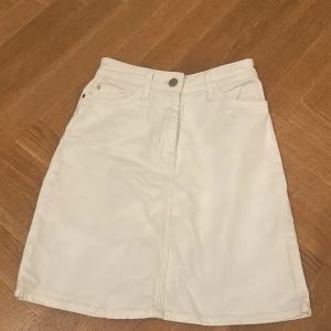 Vit kortkjol med fickor - Säljer en vit kortkjol med klassisk jeanslook och fickor fram. Kjolen har knapp och dragkedja i midjan samt bälteshällor. Perfekt för dig som gillar enkel och clean stil. Passar till sneakers eller boots.