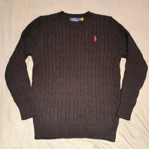 Svart kabelstickad tröja Polo Ralph Lauren - Svart kabelstickad tröja från Polo Ralph Lauren med klassisk rund halsringning och röd broderad logga på bröstet. Tröjan har långa ärmar och ribbade muddar för en snygg passform. Perfekt för dig som gillar stilrena och tidlösa st.