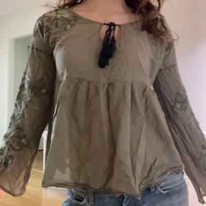 Olivgrön blus med broderade ärmar - Säljer en olivgrön blus med broderade detaljer på ärmarna och en snygg knytning vid halsen. 