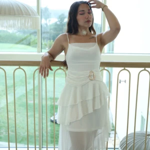 Vit långklänning med volanger och bälte - Superfin vit långklänning med smala axelband, volangdetaljer och omlottkänsla i kjolen. Klänningen har ett dekorativt bälte med guldfärgat spänne i midjan och är gjord i ett lätt, flowy material som ger en romantisk vibe. Perfekt för sommarens speciella tillfällen!