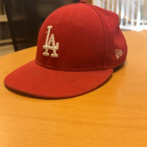 Röd LA-keps från New Era - Säljer en röd keps från New Era med LA-logga i vitt framtill. Klassisk modell med böjd skärm och broderade ventilationshål. Kepsen är tillverkad i bomull och har New Era-loggan broderad på sidan. Perfekt för dig som gillar sportig streetstyle. 7 1/4 (57,7cm)