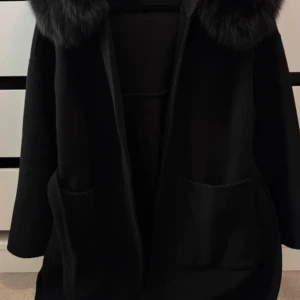 Svart kappa med fuskpäls och bälte - Säljer en svart kappa med stor krage i äktapäls och praktiska fickor framtill. Jackan har ett bälte i midjan för snygg passform och är långärmad. Perfekt för dig som vill ha en stilren och varm look under höst och vinter.