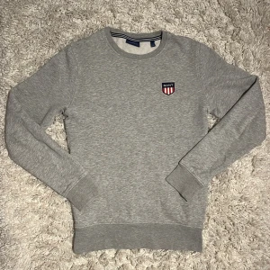Grå sweatshirt från GANT med logo - Klassisk grå sweatshirt från GANT i bomull med rund hals och broderad GANT-logo på bröstet. Tröjan är i nytt skick och har storlek S. 