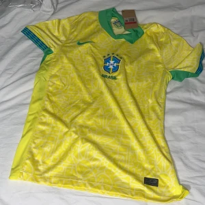 Brasilien Nike fotbollströja gul/grön - Säljer en officiell Brasilien fotbollströja från Nike i gult med gröna detaljer och blå Nike-logga. Tröjan har korta ärmar, mönstrad framsida och broderat Brasilien-emblem på bröstet. Perfekt för dig som vill stötta landslaget med stil.