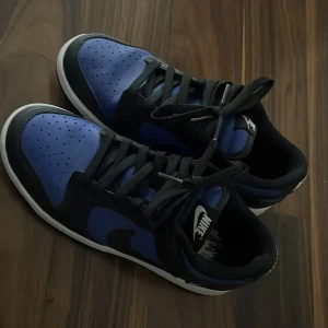 Nike Dunk Low blå och svarta sneakers - Säljer ett par Nike Dunk Low sneakers i blått och svart med vit sula. Klassisk design med svart swoosh, snörning och rund tå. Ovandelen är i syntet och skinn, med perforerade detaljer på tån för extra stil. Perfekta för dig som gillar streetwear och vill sticka ut.