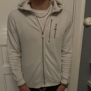 Vit hoodie från Sail Racing - Säljer en stilren vit hoodie från Sail Racing med dragkedja framtill och en extra bröstficka med zip. Tröjan har huva med snörning och diskret logga på bröstet. Perfekt för dig som gillar en clean och sportig look.