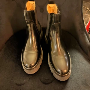 Svarta chelsea boots med grov sula från Gant - Snygga svarta chelsea boots i gummi  med grov, räfflad sula och guldfärgade detaljer baktill. Bootsens insida är beige och de har elastiska paneler på sidorna för enkel påtagning. Perfekta för dig som vill ha en stilren men edgy look.       Har tyvärr inte kvar originalkartongen, men aldrig använda utan endast provat dem.                   Nypris 1900kr