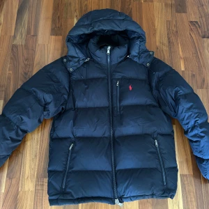 Mörkblå dunjacka från Ralph Lauren - Snygg mörkblå dunjacka från Ralph Lauren med huva och dragkedja framtill. Jackan har tre fickor med dragkedja och den klassiska röda Polo-loggan på bröstet. Använd i ett år, inga defekter eller hål. Perfekt till vintern.