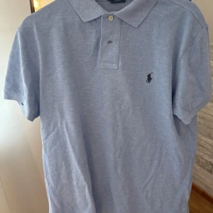 Ljusblå pikétröja från Ralph Lauren - Säljer en klassisk ljusblå pikétröja från Ralph Lauren med korta ärmar och broderad logga på bröstet. Tröjan har krage och knäppning med två knappar. Perfekt för en clean och stilren look. Slim fit storlek L passar M