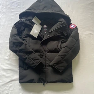 Svart dunjacka från Canada Goose XS - Svart dunjacka från Canada Goose med huva och klassisk logga på ärmen. Jackan har dragkedja och knappar framtill, två sidofickor och en stilren, enkel design. Perfekt för kalla dagar och riktigt snygg till vintern.