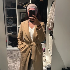 Beige kappa med bälte och knappar - Snygg beige kappa med klassisk krage, knappar framtill och matchande bälte i midjan. Lång modell med stilren look, perfekt för dig som gillar en clean och tidlös stil. Kappan har långa ärmar och är tillverkad i ett mjukt material som känns lyxigt.
