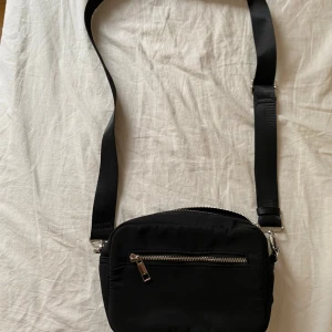 Svart axelväska med dragkedjor - En stilren svart axelväska med justerbart band och flera fack med silvriga dragkedjor. Väskan är kompakt och smidig, perfekt för att bära det viktigaste. Tillverkad i ett mjukt syntetmaterial som känns lätt och bekvämt att ha på sig.
