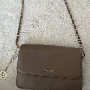 Vintage Beige axelväska från DKNY i skinn - Snygg vintage beige axelväska från DKNY i slätt skinn med guldfärgad kedja och läderdetaljer på axelremmen. Väskan har ett stilrent DKNY-emblem framtill och flera fack inuti, inklusive dragkedjefack. Perfekt storlek för det viktigaste och en klassisk look.