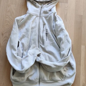 Vit hoodie från Sail Racing med huva - Vit hoodie från Sail Racing med dragkedja framtill, huva och en bröstficka med logga. Insidan är fodrad med mjuk fleece för extra komfort. Perfekt för kyliga dagar och har en clean, sportig look.