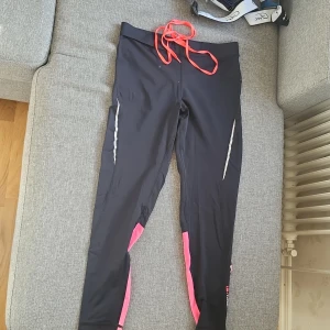 Svarta träningsbyxor från Tenson - Svarta träningsbyxor från Tenson med rosa detaljer vid bensluten och reflexränder på sidorna. Byxorna har snörning i midjan och en liten ficka med dragkedja baktill. Perfekta för löpning eller annan träning.