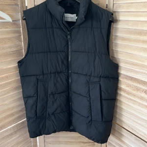 Svart dunväst från Calvin Klein M - Svart quiltad dunväst från Calvin Klein med hög krage och dragkedja framtill. Västen har två stora fickor på sidorna och diskret logga på ryggen. Perfekt för lager-på-lager och enkel att matcha med olika outfits.